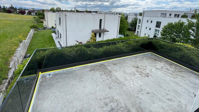 Flachdachterrasse mit grauer Abdichtungsfläche und Glasgeländer, Blick auf Wohnhäuser.