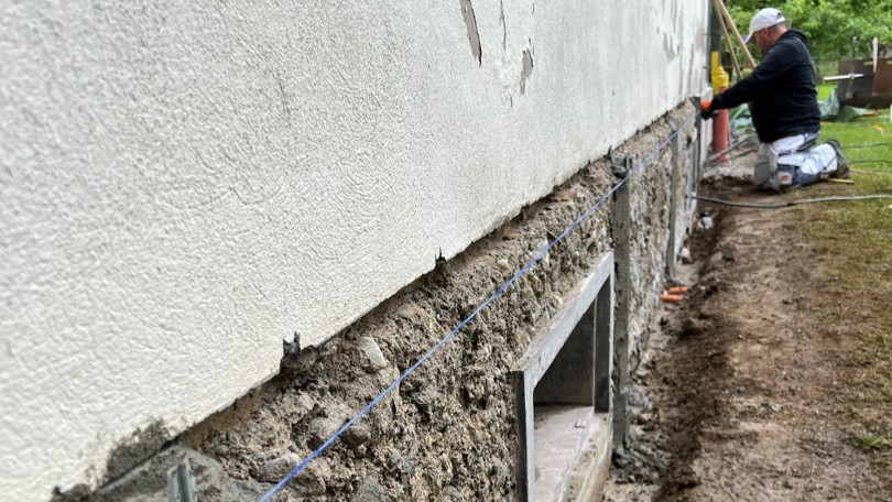 Arbeiter glättet graue Masse an Sockelbereich eines Gebäudes, Erdgraben daneben.