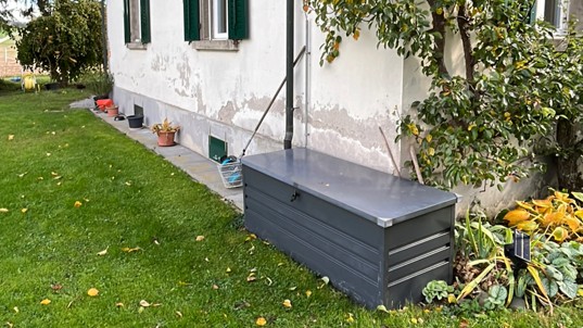 Hausfassade mit abplatzendem Putz neben Rasen und grauer Box am Sockel.
