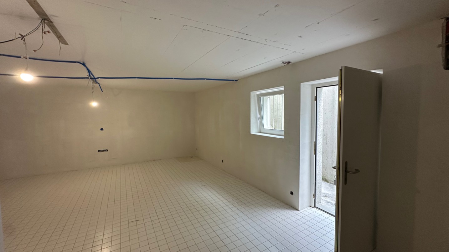 Leerer Kellerraum mit Rasterboden, offenem Türzugang und sichtbaren Deckenleitungen