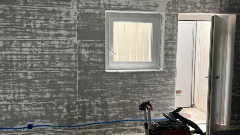 Graue Wandfläche mit Fenster, offener Tür und blauer Leitung am Boden
