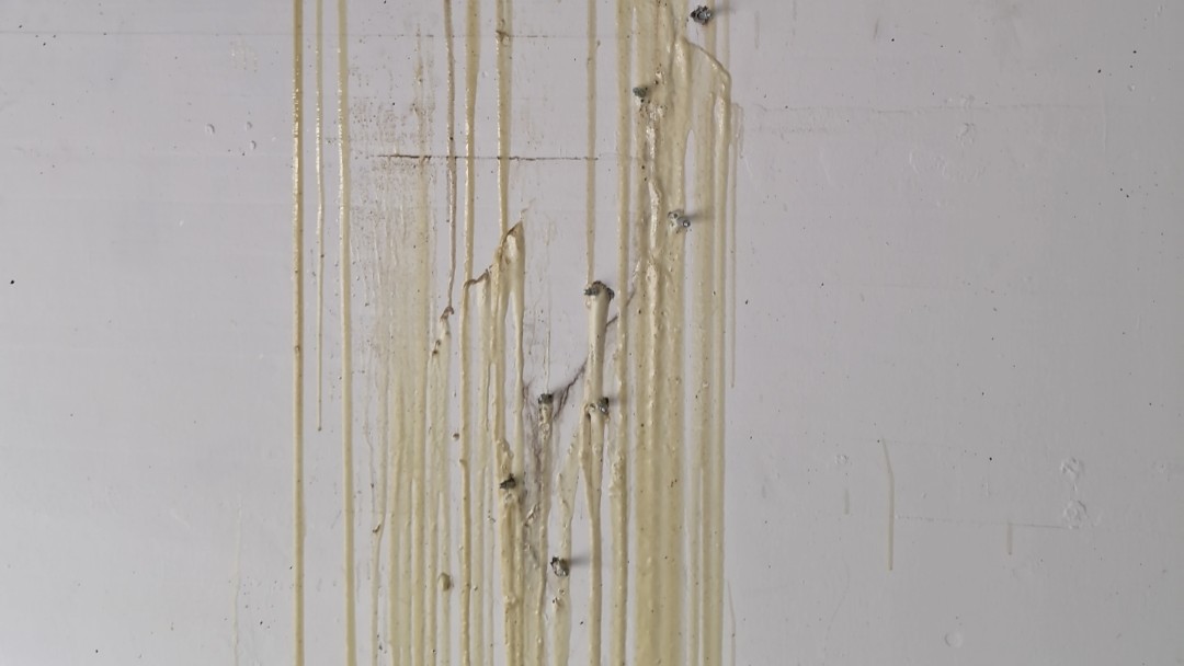 Cremefarbene Masse läuft in senkrechten Bahnen über weiße Wand, aus der kleine Metallstifte herausragen.