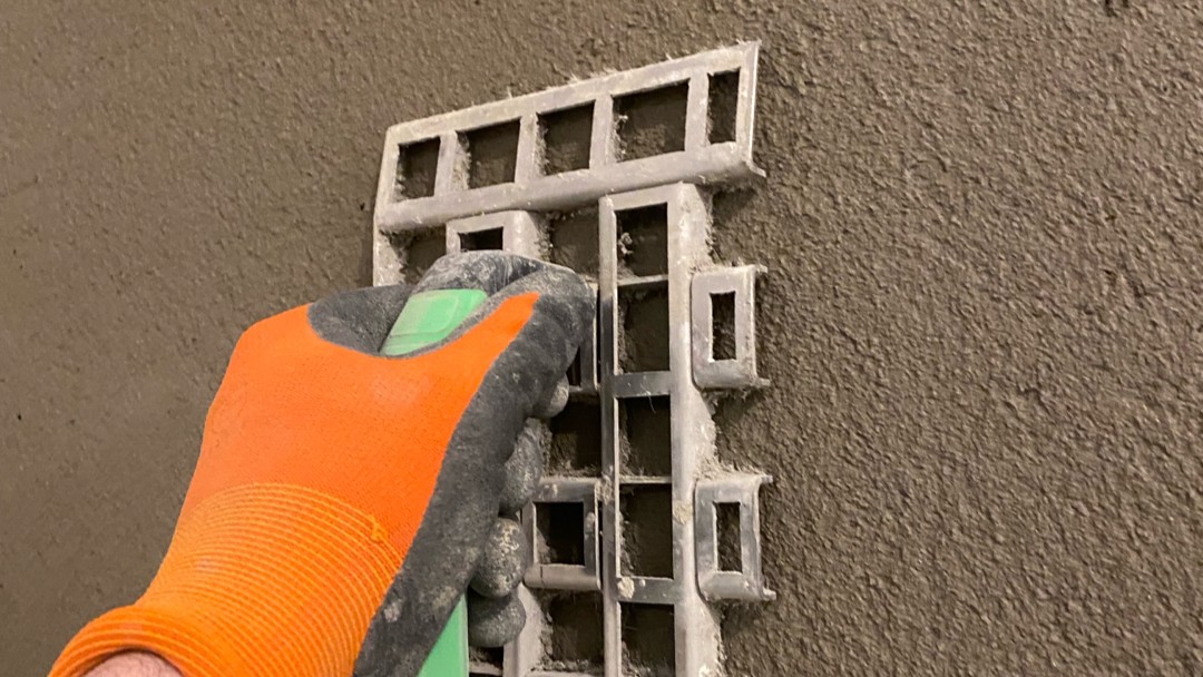 Hand mit orangefarbenem Handschuh setzt Kunststoffprofil in graue Masse an Wand