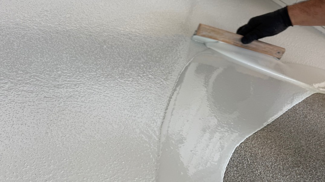 Handschuhte Hand zieht graue Masse mit Glättkelle über strukturierte Fläche.