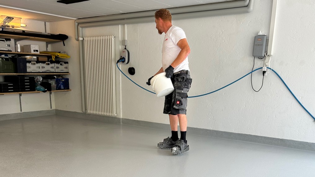 Arbeiter verteilt transparente Flüssigkeit auf grauem Boden in Raum.
