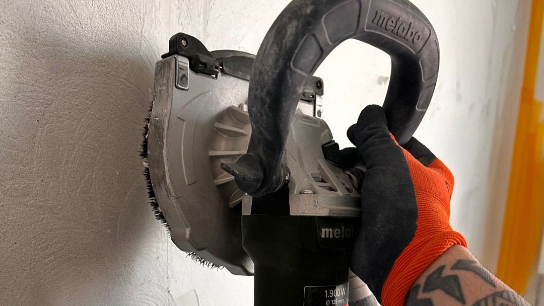 Hand führt Schleifmaschine mit Bürstenkranz an rauer Wand entlang