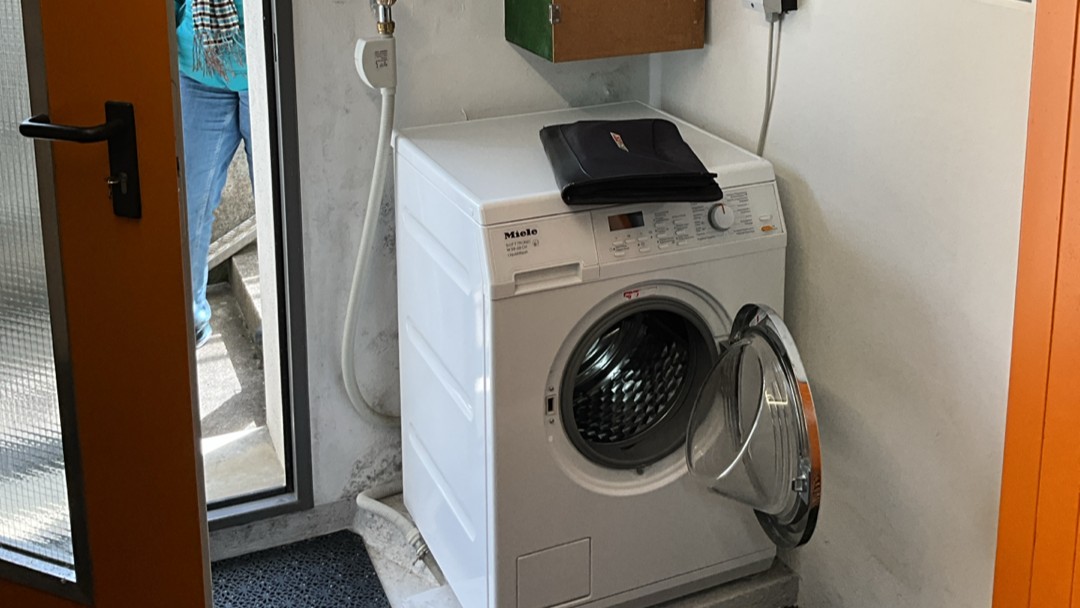 Weiße Waschmaschine mit offener Tür steht vor grauer Wand, daneben Zulaufschlauch.
