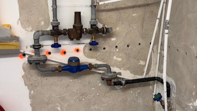 Blauer Wasserzähler und Messing-Druckminderer an Metallrohren an grauer Wand.