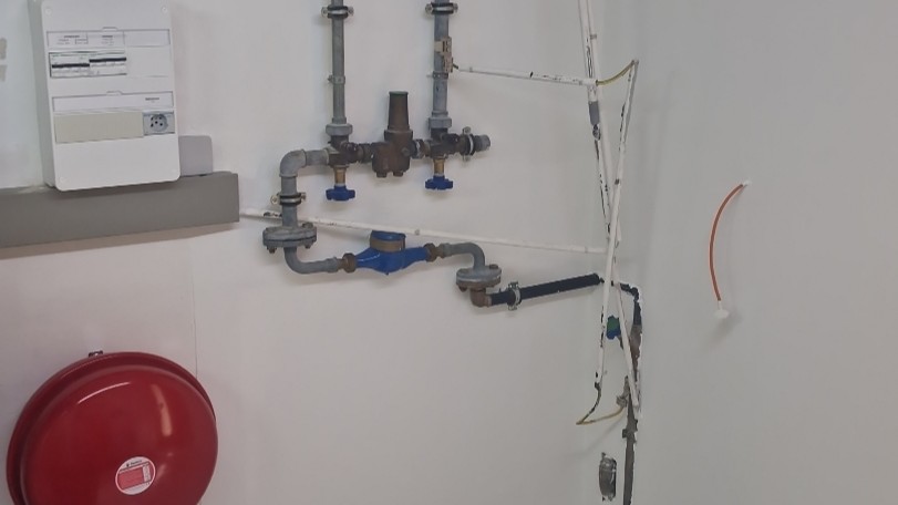 Metallrohre mit Druckminderer und blauem Wasserzähler, rotes Ausdehnungsgefäß an weißer Wand