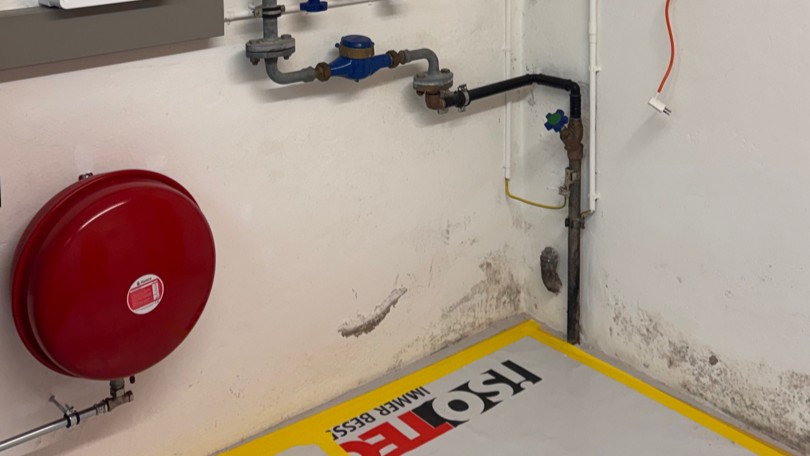 Rotes Ausdehnungsgefäß und Rohrleitungen an Wand mit dunklen Feuchtflecken, Boden mit ISOTEC-Markierung