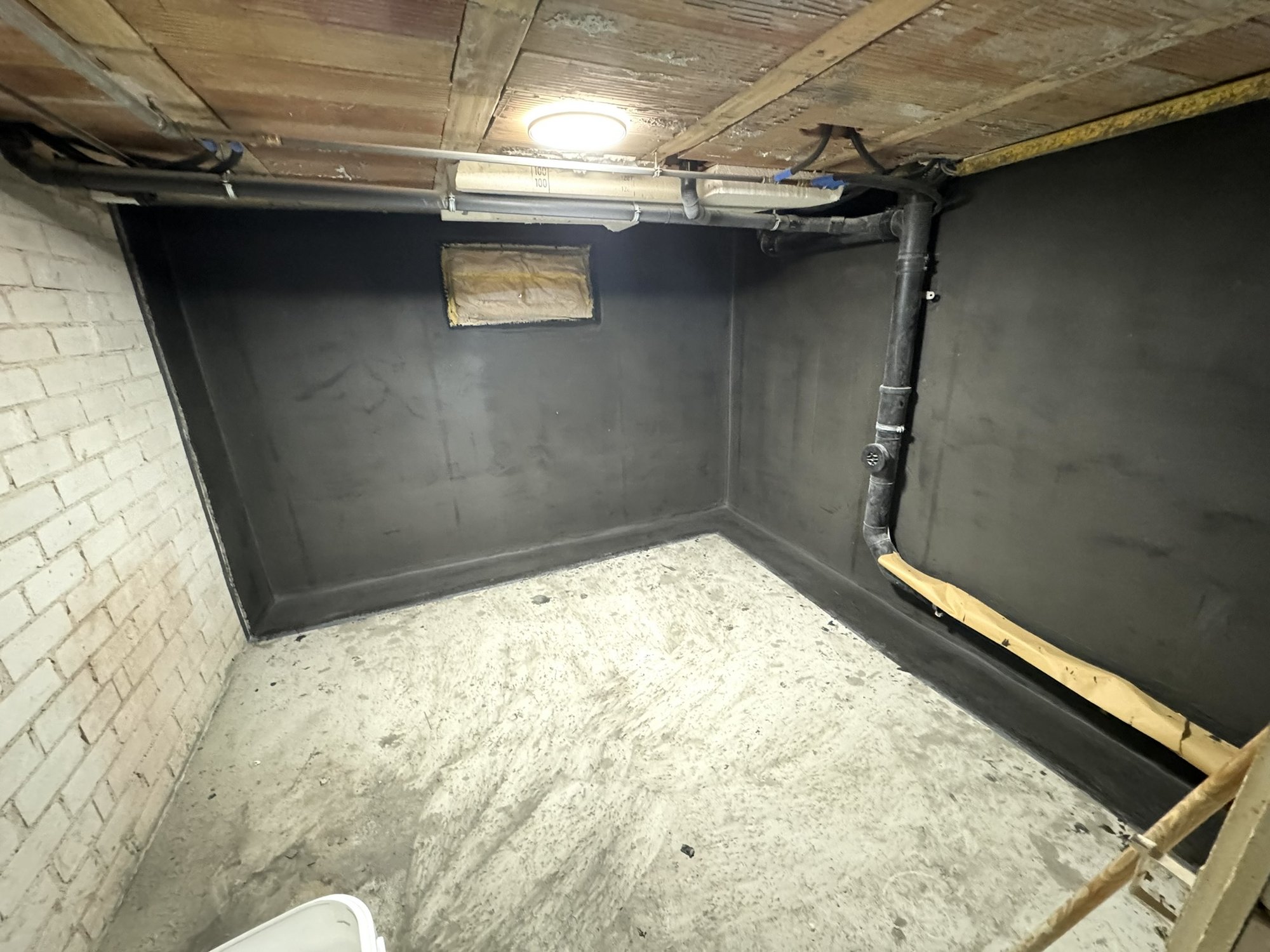 Kellerraum mit schwarzer Masse beschichteten Wänden, sichtbaren Rohren und kleinem Fenster