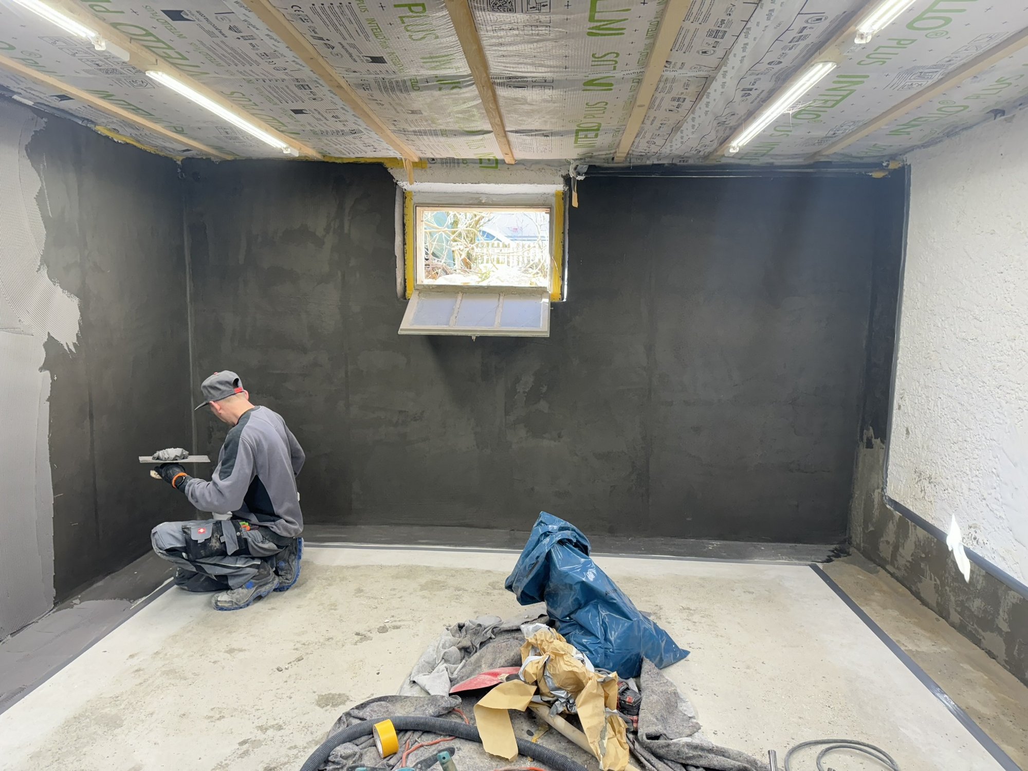 Arbeiter glättet schwarze Masse an Kellerwand unter gedämmter Decke.