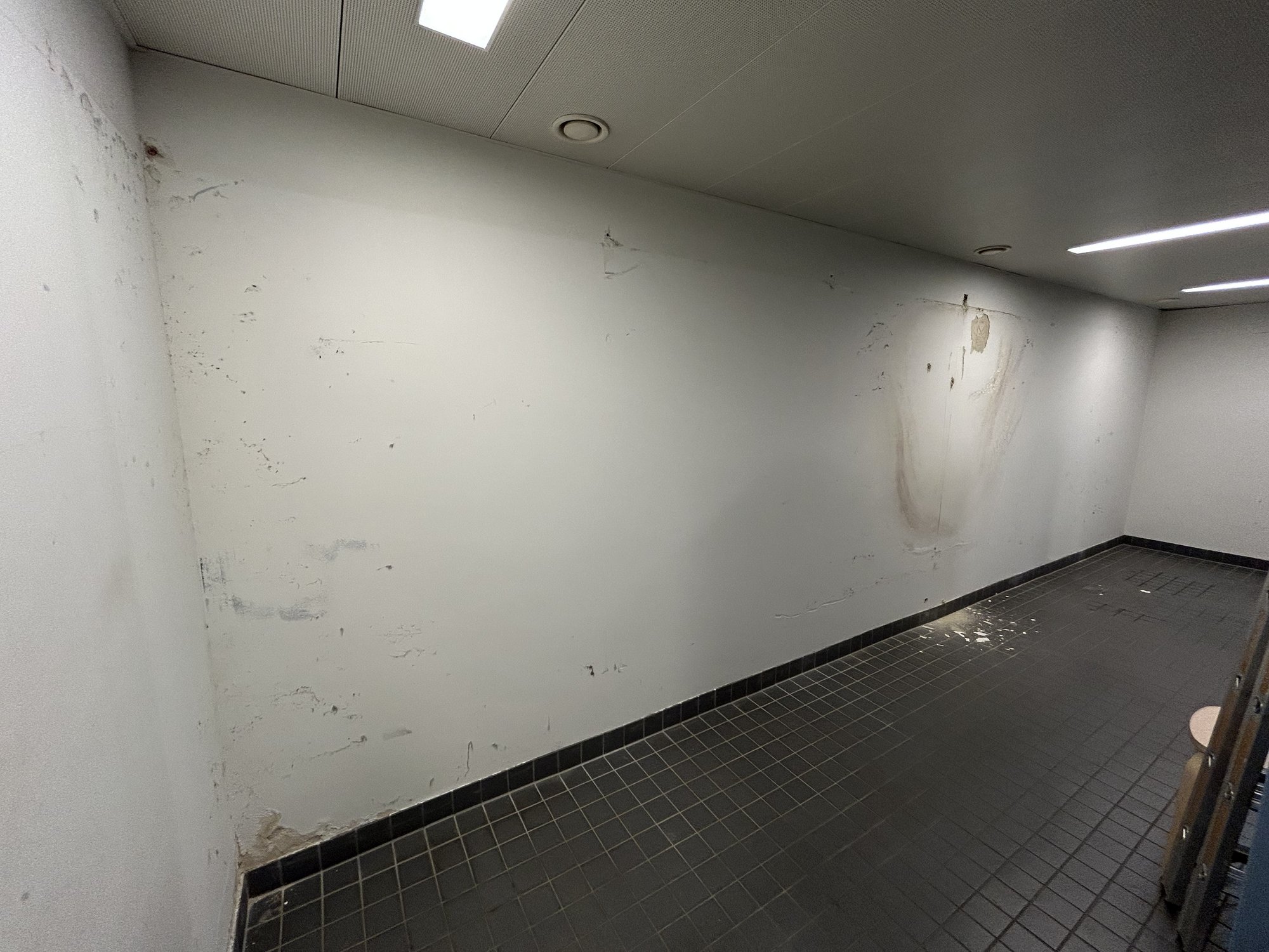 Weißer Raum mit Fliesenboden, Wand mit Flecken, Rissen und abplatzender Beschichtung.