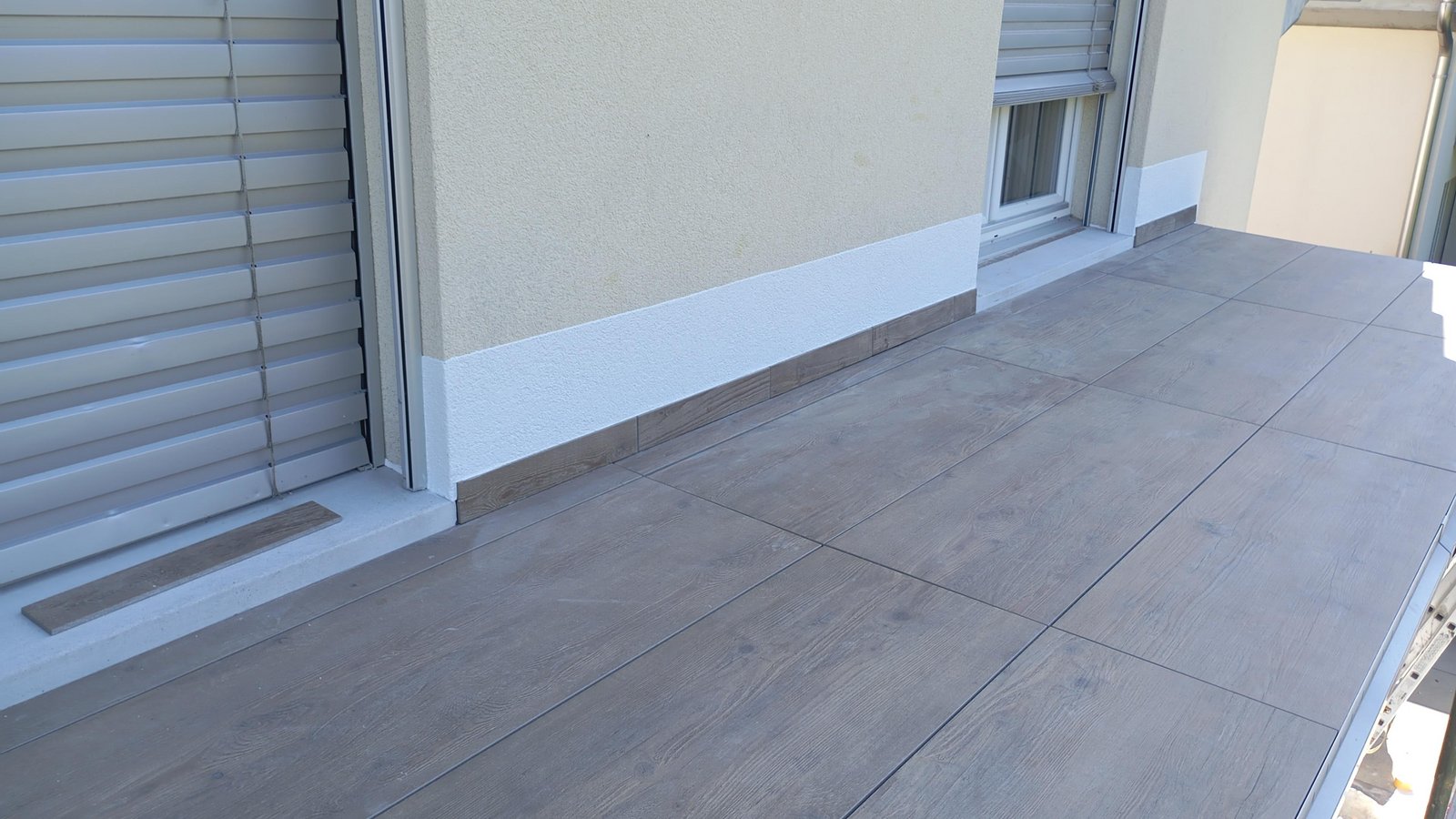 Balkon mit grauen Bodenfliesen, gelber Fassade und Fenster mit Rollladen